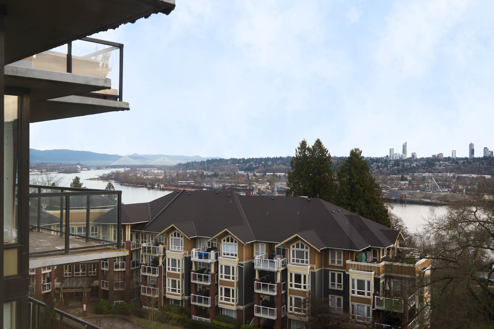 View from a balcony overlooking residential buildings, a river, and a distant city skyline under a partly cloudy sky.
