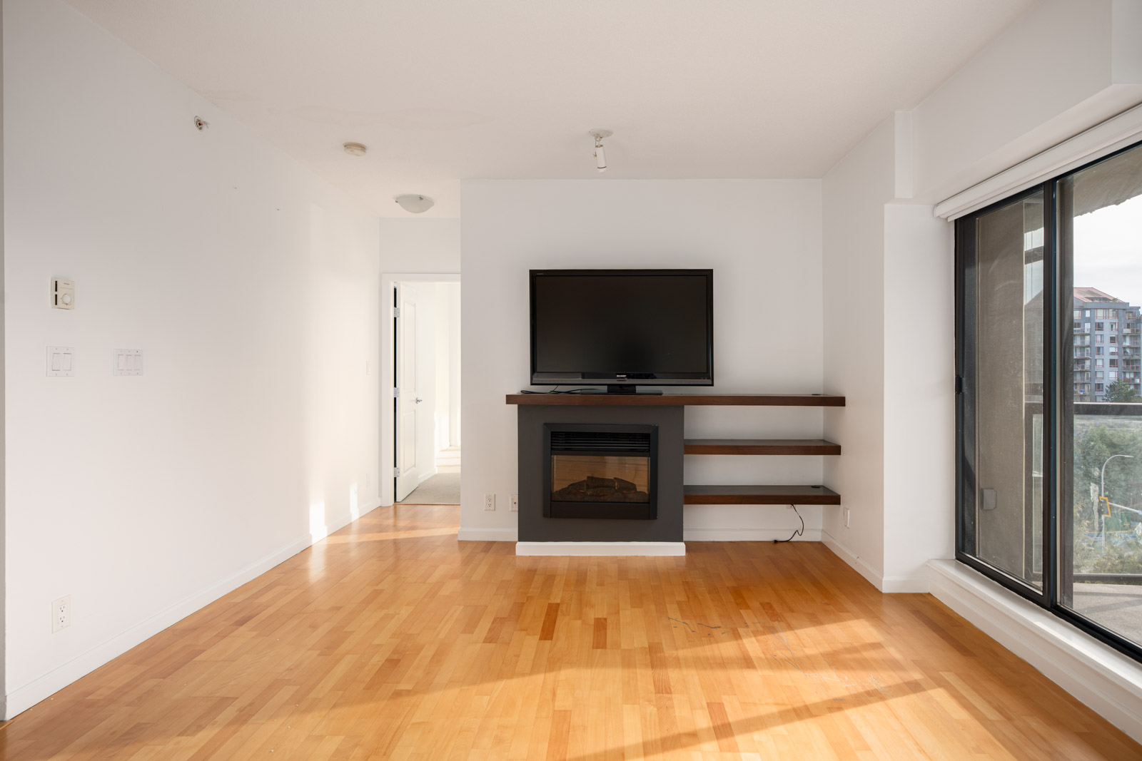 A modern living room with light wood flooring, a wall-mounted TV above a fireplace, floating shelves, and a large window with a view of nearby buildings.