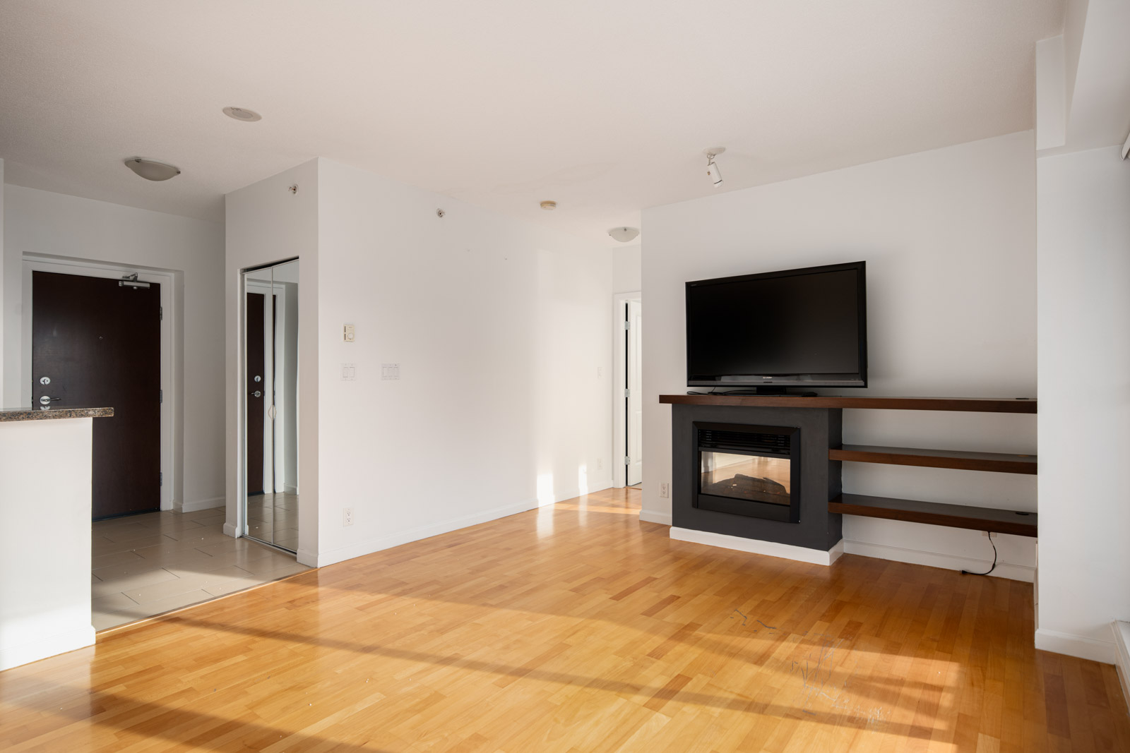 Bright living room with hardwood floor, a wall-mounted TV above a fireplace, built-in shelves, and a view of an entrance hallway with a mirrored closet door.