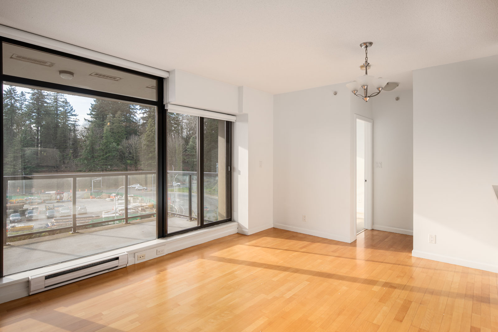 Bright, unfurnished apartment room with light wood flooring, large windows, balcony access, white walls, and a ceiling light fixture. Construction site and trees visible outside.