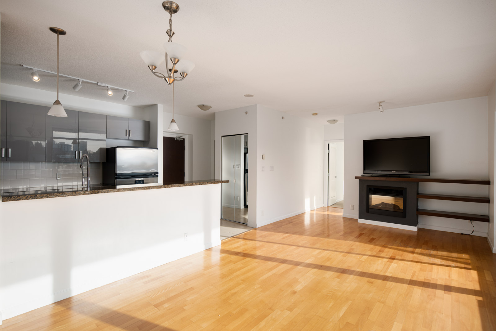 Bright, modern open-concept kitchen and living area with hardwood floors, white walls, stainless steel appliances, wall-mounted TV, fireplace, and large windows letting in natural light.