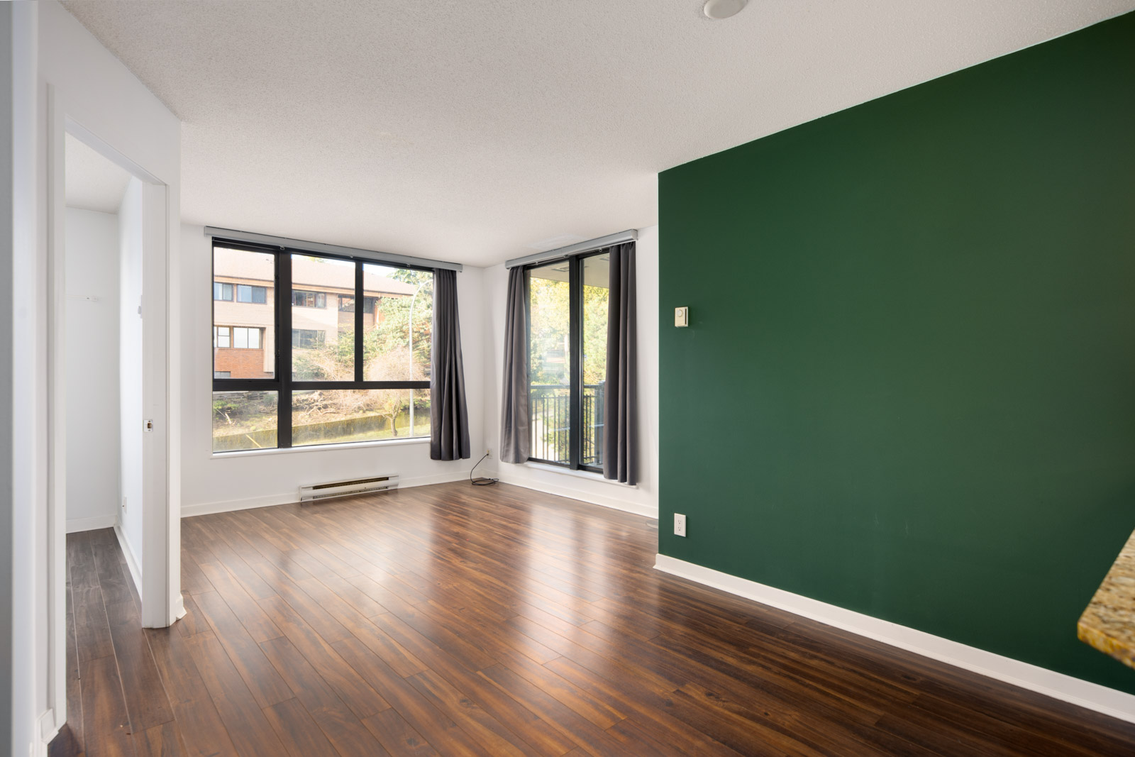Empty living room with dark wood floors, a green accent wall, large windows, and a view of outdoor greenery and a neighboring building.