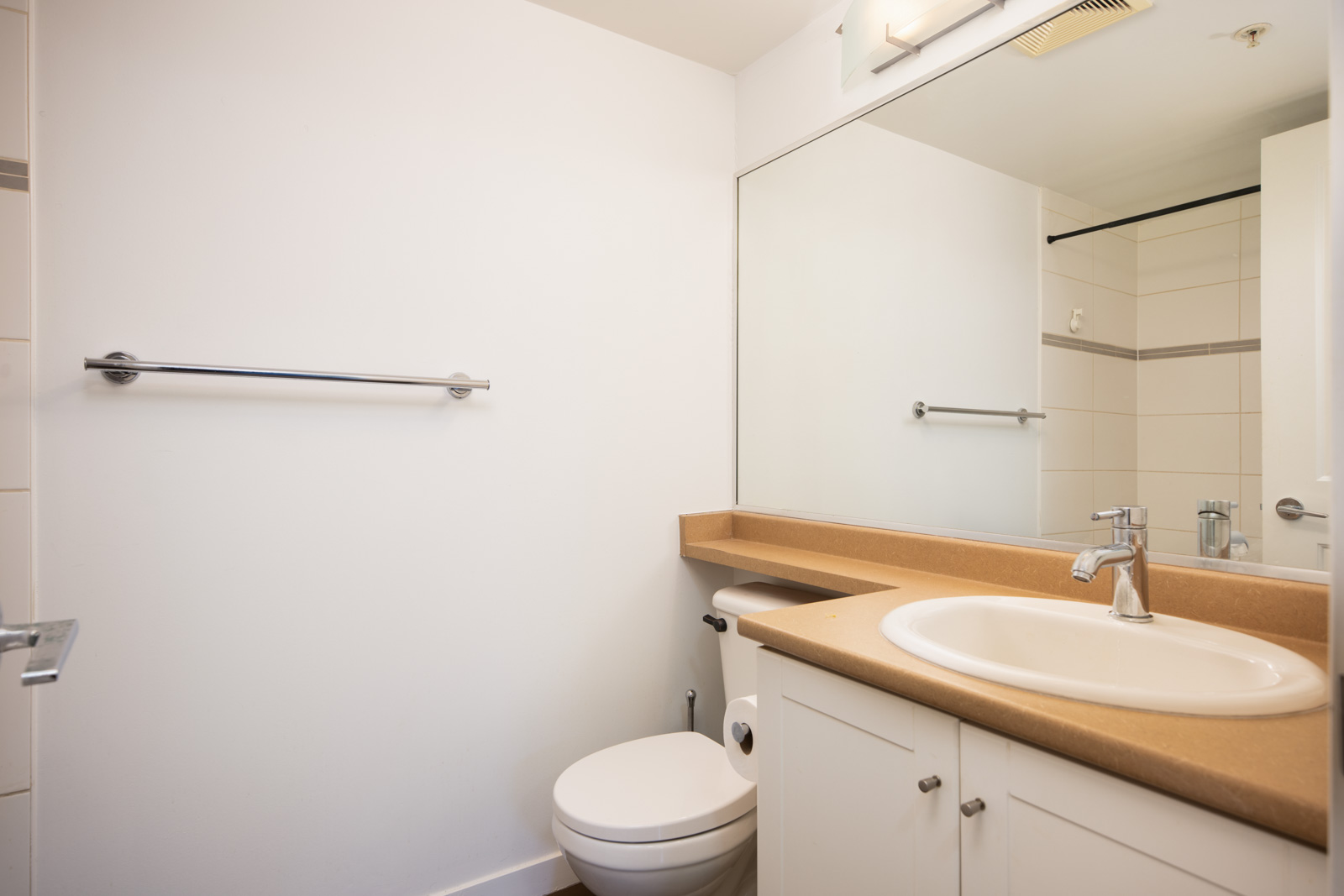 A bathroom with a beige countertop, white sink, cabinet, toilet, large wall mirror, towel bar, and white tiled walls.