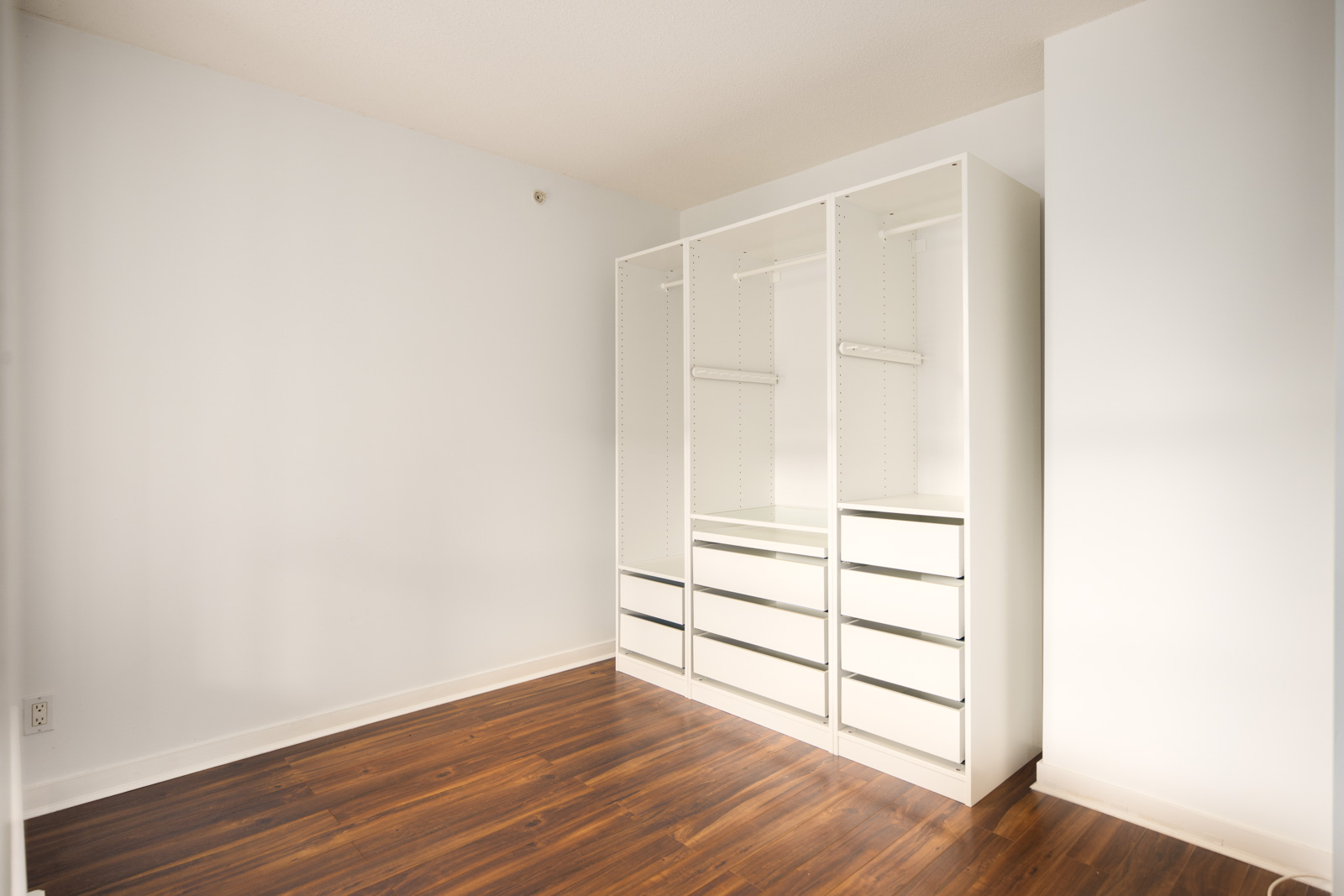 A minimalist closet with white shelving units and drawers is placed against a white wall in a room with wood flooring.