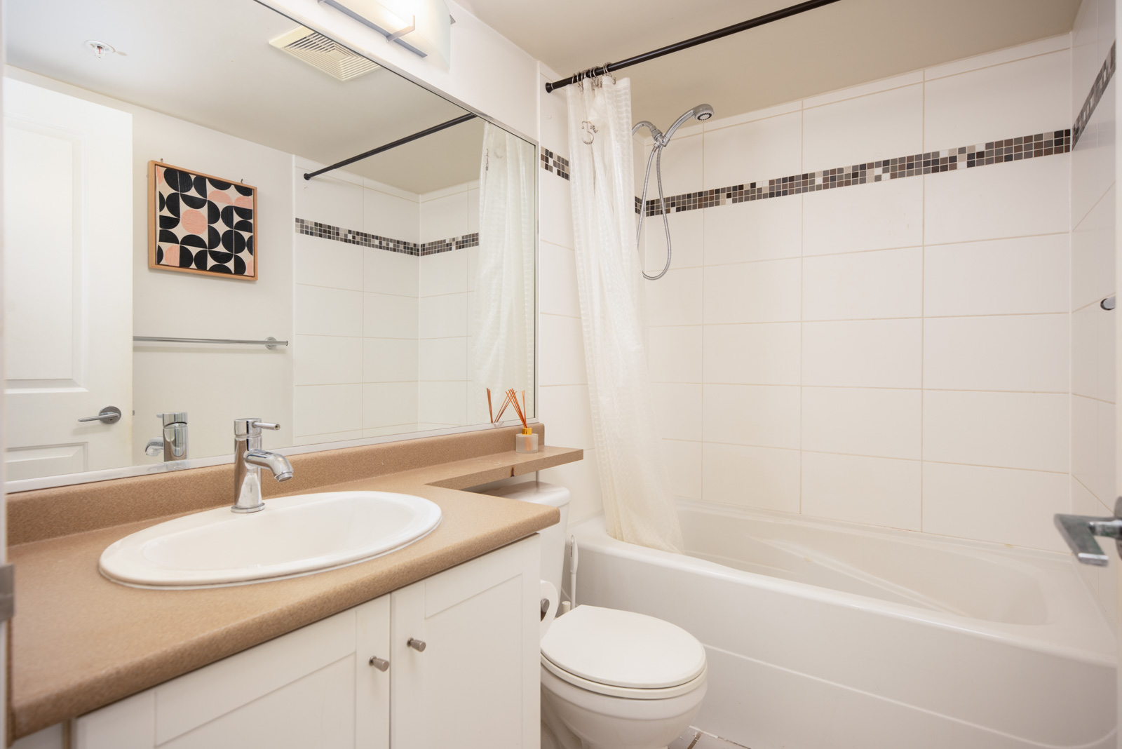 Modern bathroom with a white sink, countertop, toilet, bathtub, shower curtain, wall tiles, large mirror, and abstract artwork on the wall.