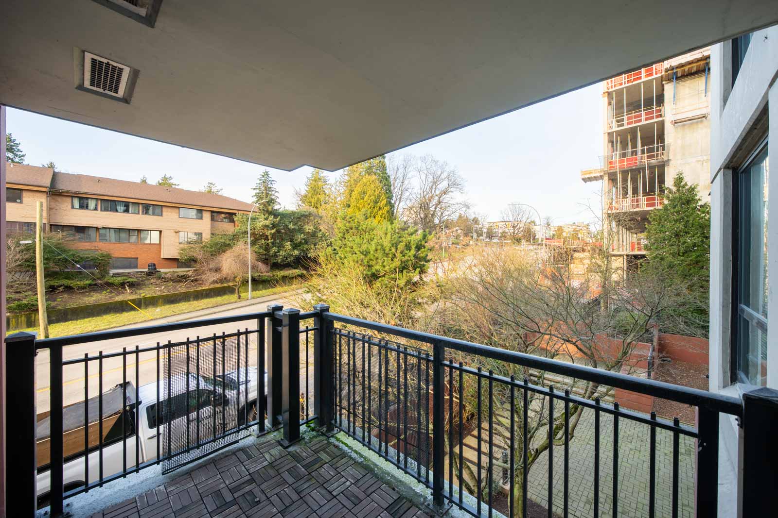 View from a balcony with black metal railings, overlooking a quiet street, residential buildings, trees, and a nearby construction site.
