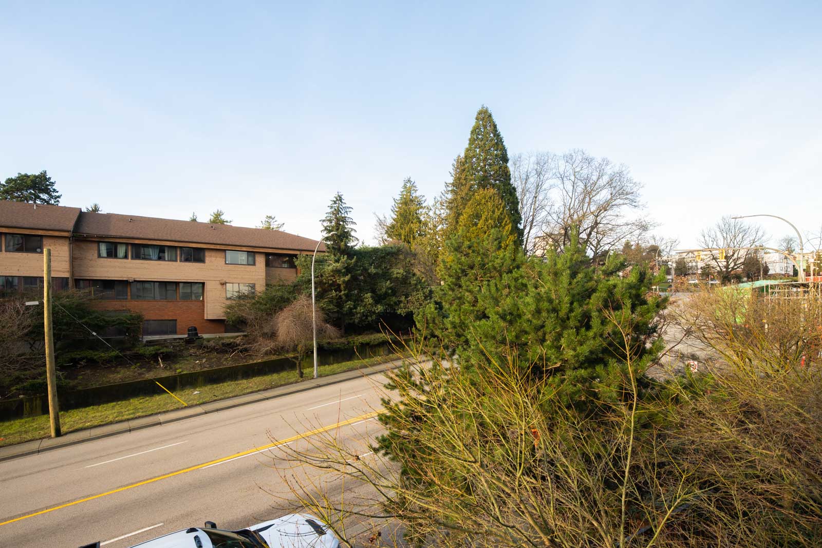 A quiet residential street with trees and shrubs, a brick apartment building on the left, and a clear blue sky overhead.