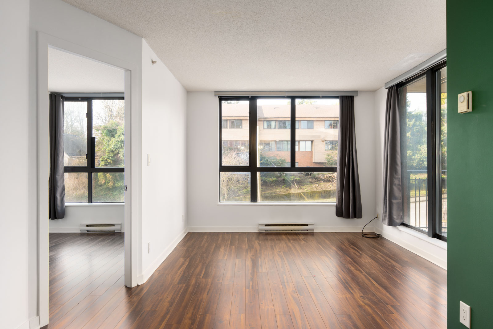 Empty, unfurnished apartment room with large windows, hardwood floors, white walls, and gray curtains; neighboring building visible outside.