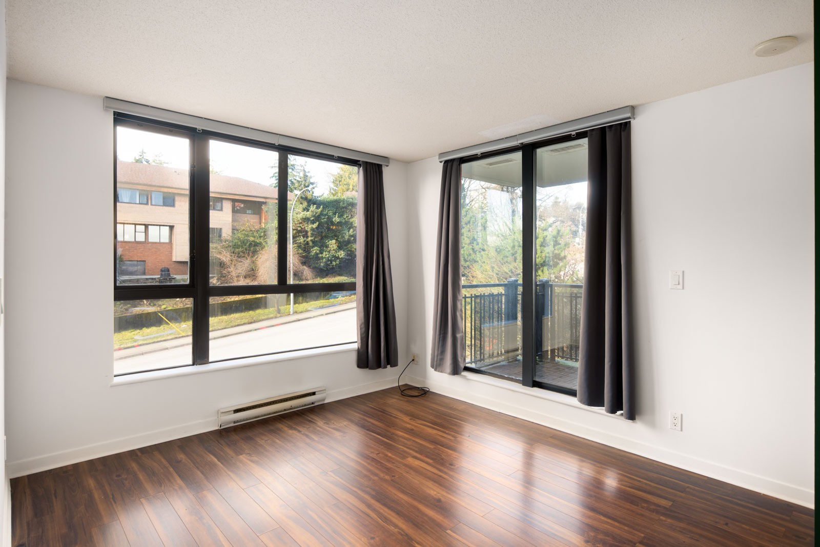 Empty room with wood flooring, large corner windows with black curtains, a glass door leading to a balcony, and white walls.