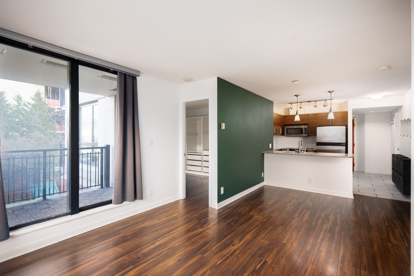 Modern apartment interior with wood flooring, a green accent wall, open kitchen with stainless steel appliances, and large sliding glass doors leading to a balcony.