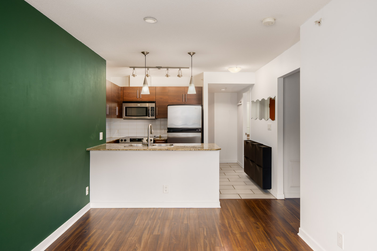 Modern kitchen with stainless steel appliances, granite countertop, wooden cabinets, and a green accent wall, adjacent to a hallway with dark wood flooring.