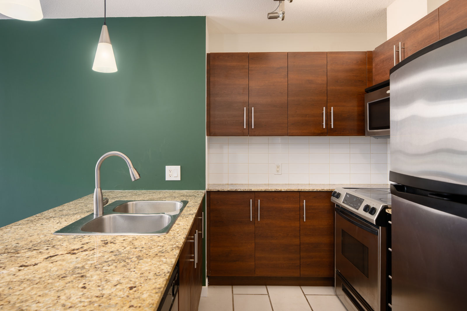 Modern kitchen with dark wood cabinets, granite countertop, stainless steel appliances, double sink, green accent wall, and tile backsplash.