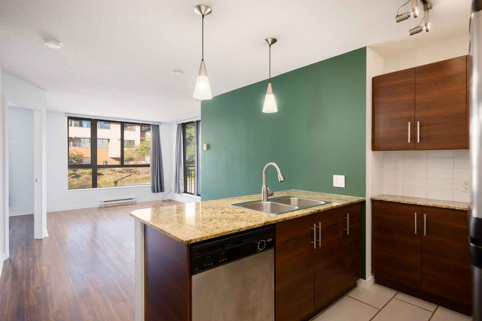 Modern apartment kitchen with granite countertops, dark wood cabinets, pendant lights, and a green accent wall, opening to a bright living area with large windows.