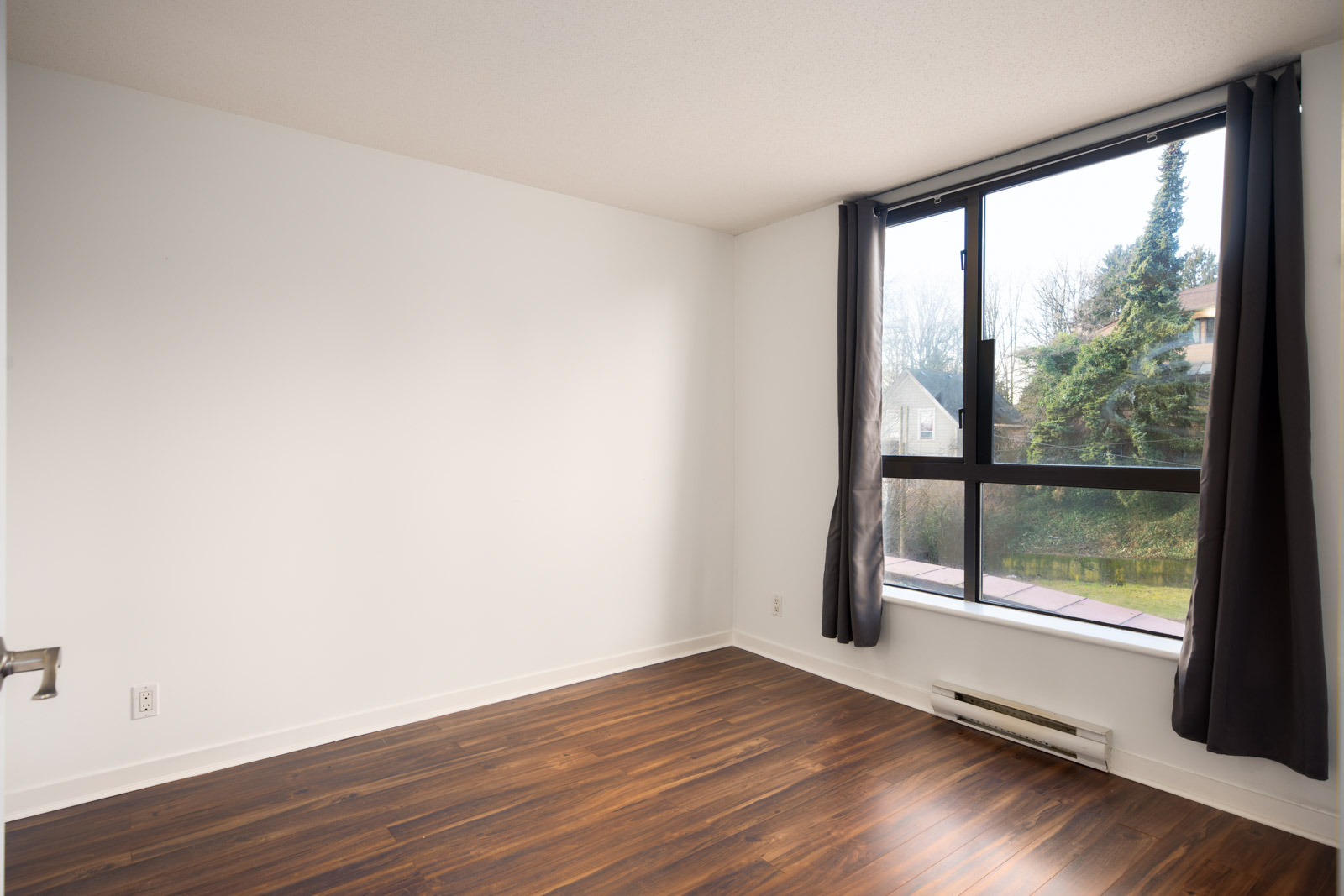 Empty room with white walls, dark wood flooring, a large window with black curtains, and baseboard heater beneath the window.