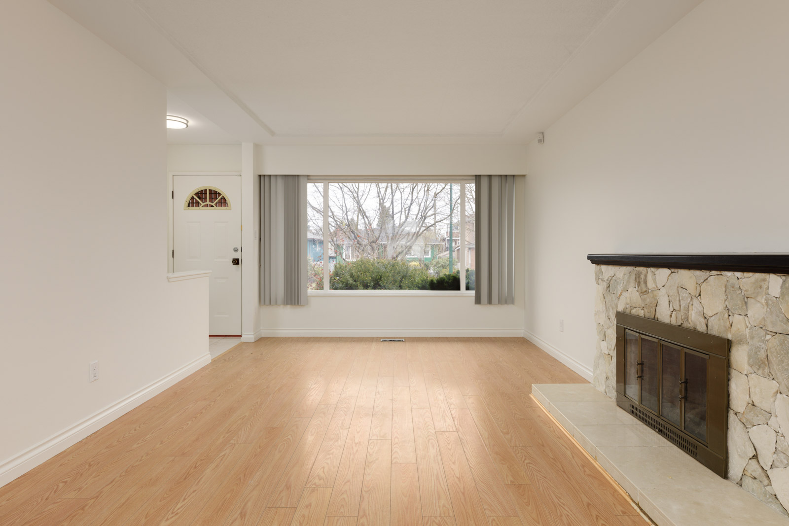 Empty living room with light wood flooring, a large front window with closed curtains, a white entrance door, and a stone fireplace with a glass screen.