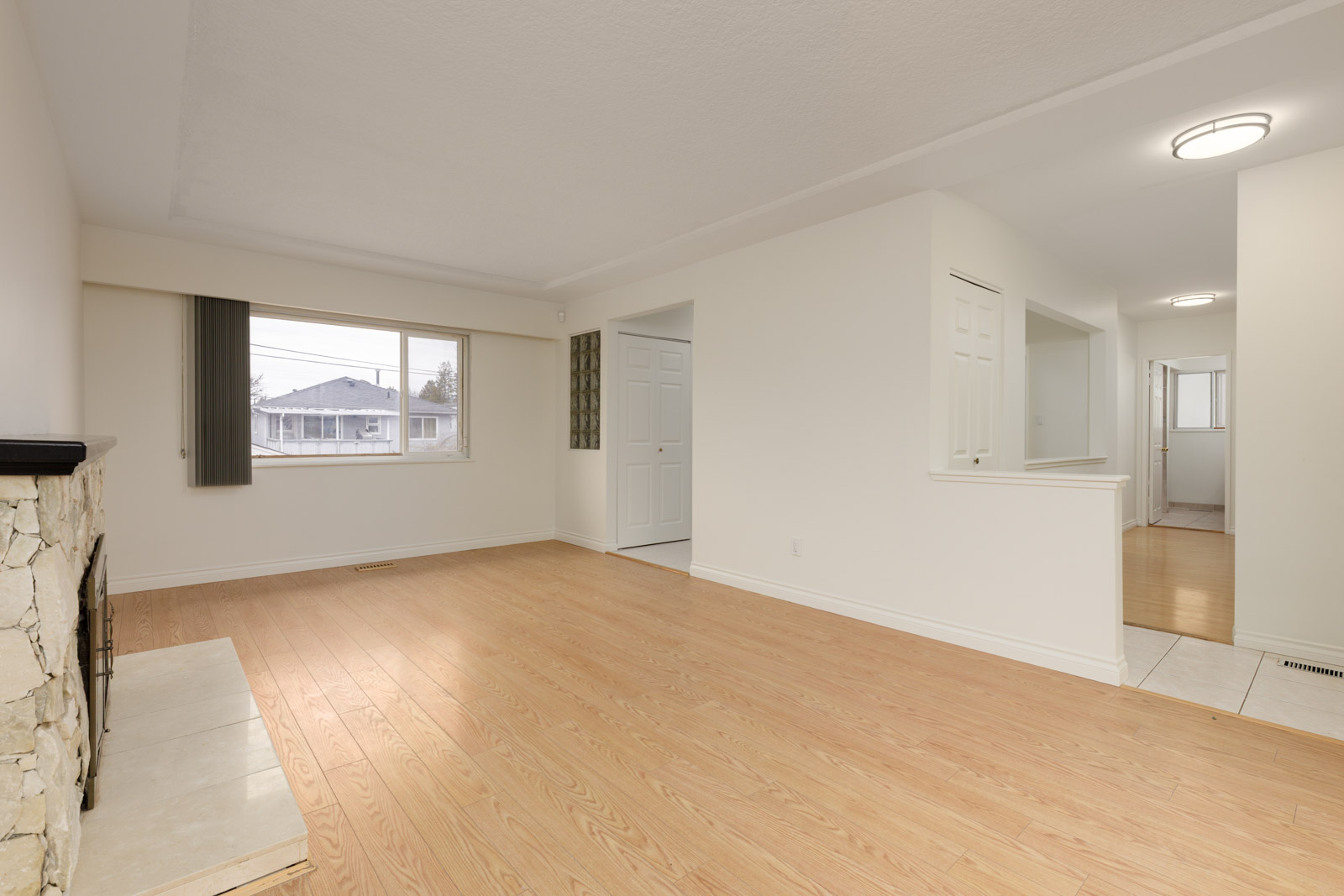 Empty living room with light wood flooring, white walls, a stone fireplace on the left, large window, and open doorway leading to a hallway and tiled area.