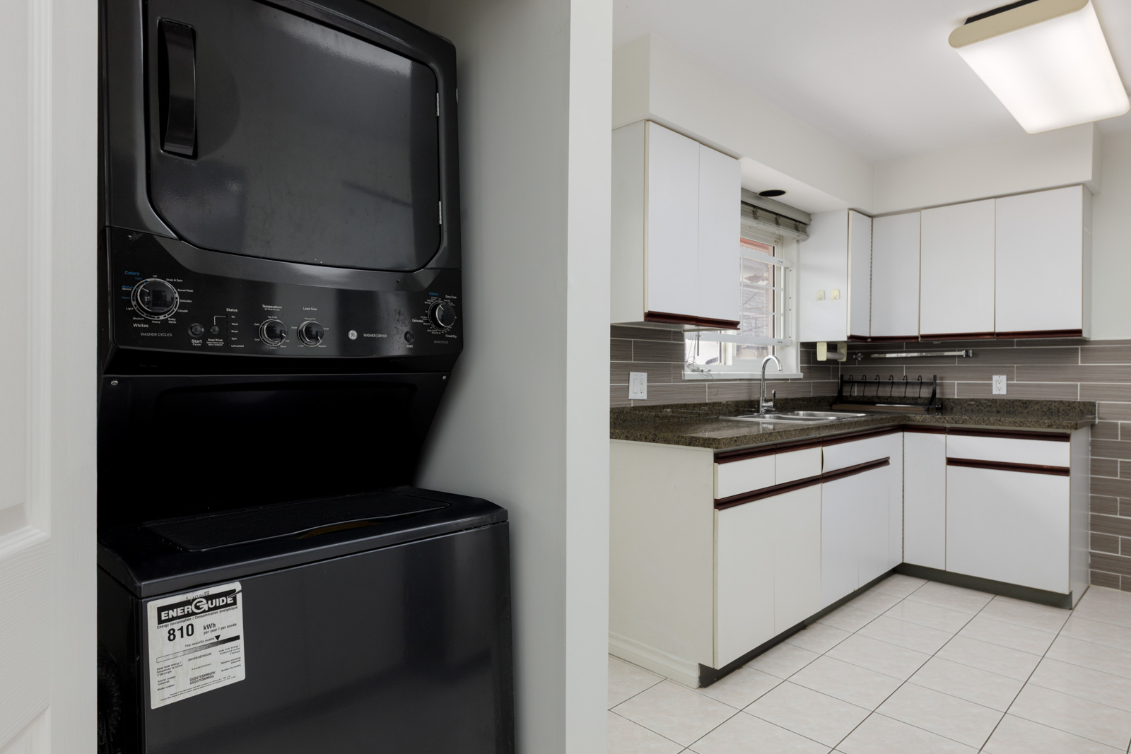 A stacked black washer and dryer unit is next to a modern kitchen with white cabinets, granite countertops, and tile flooring.