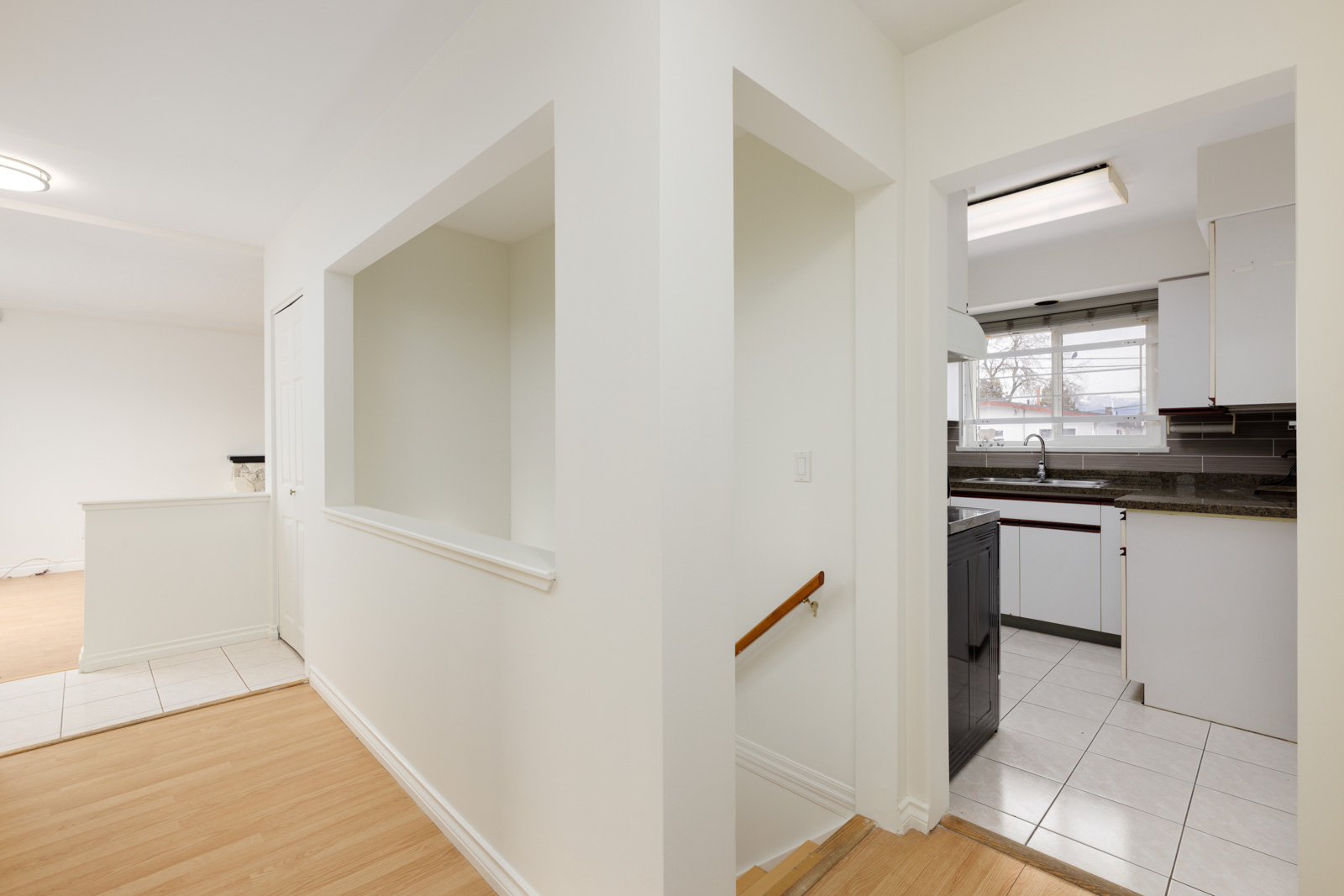 Hallway with light wood flooring leads to a stairway and a kitchen with tile floor, white cabinets, and a window above the sink. Open wall cutout connects hallway to another room.