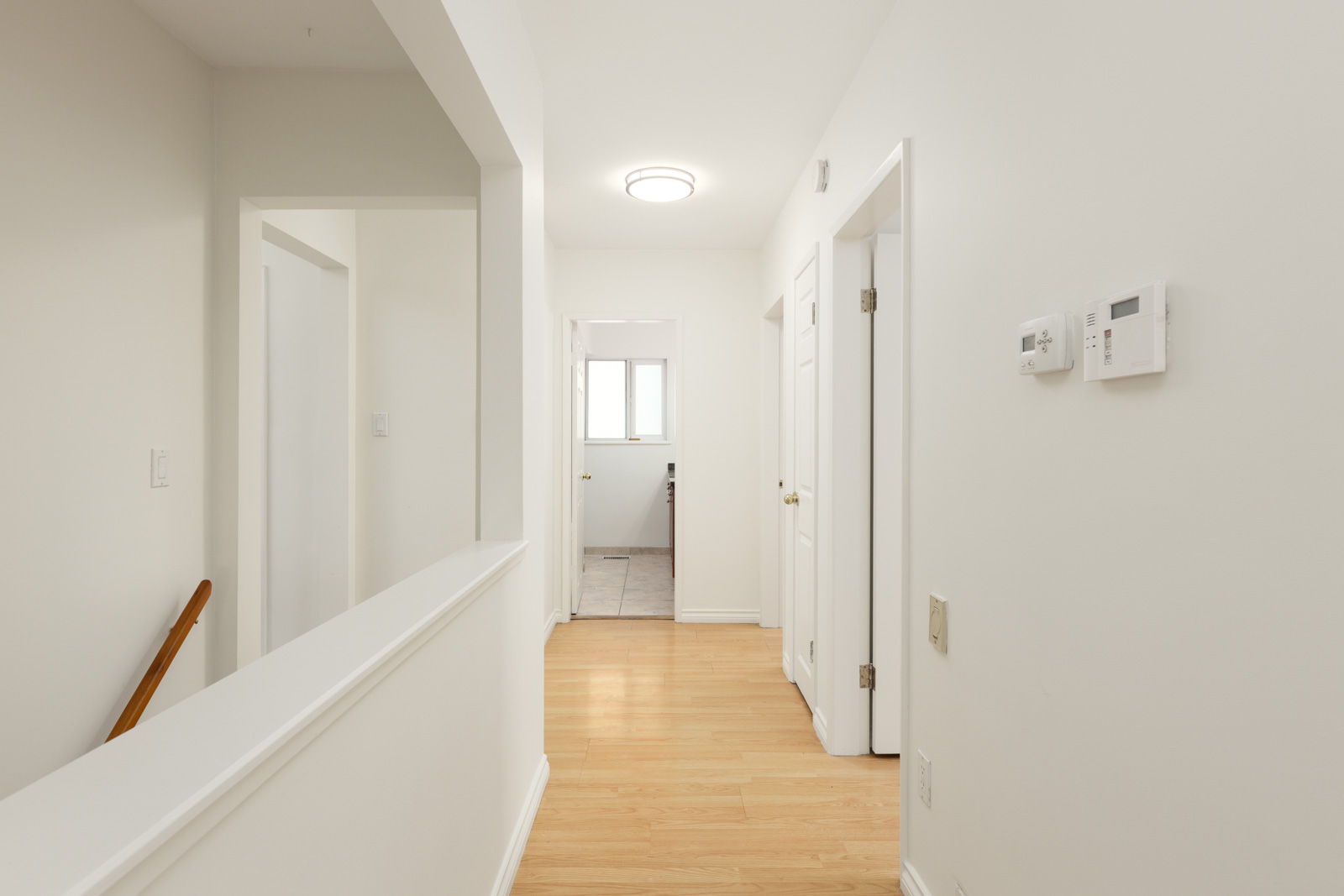 A hallway with light wood flooring, white walls, and doors leading to adjacent rooms. A thermostat and light switches are mounted on the right wall. Natural light enters from a far window.