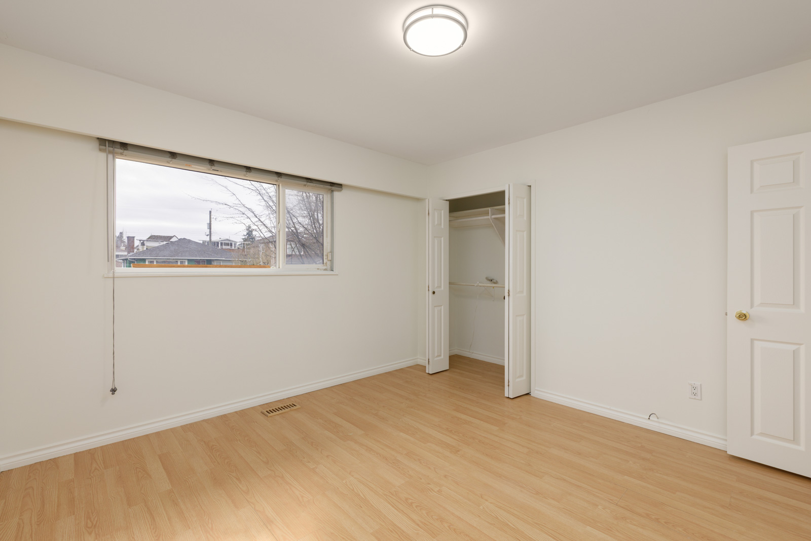 Empty room with light-colored walls, wood flooring, a large window, a closet with double doors, and a closed door on the right. Ceiling light fixture is turned on.