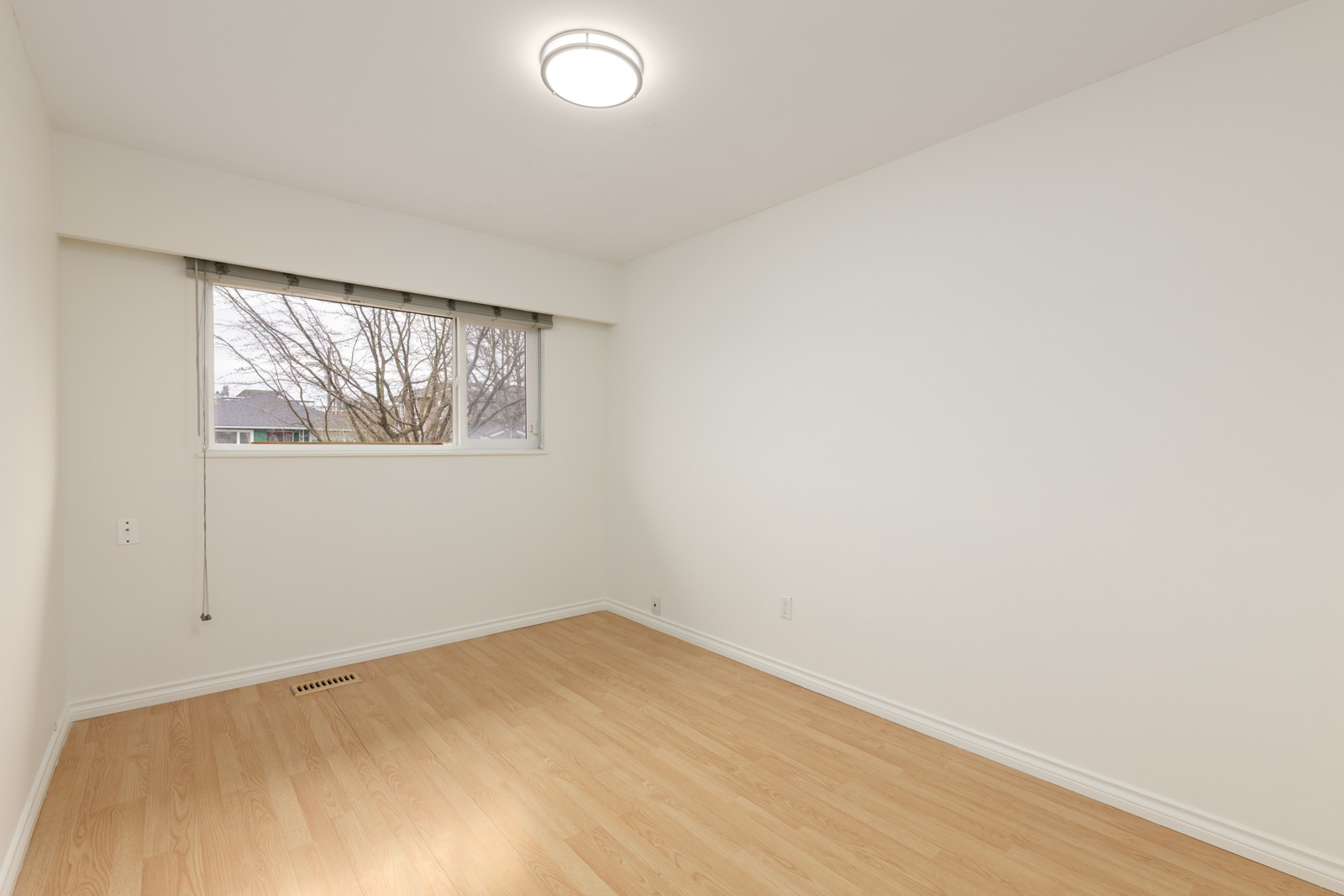 Empty room with light wood flooring, white walls, a ceiling light, and a window with a view of tree branches outside.