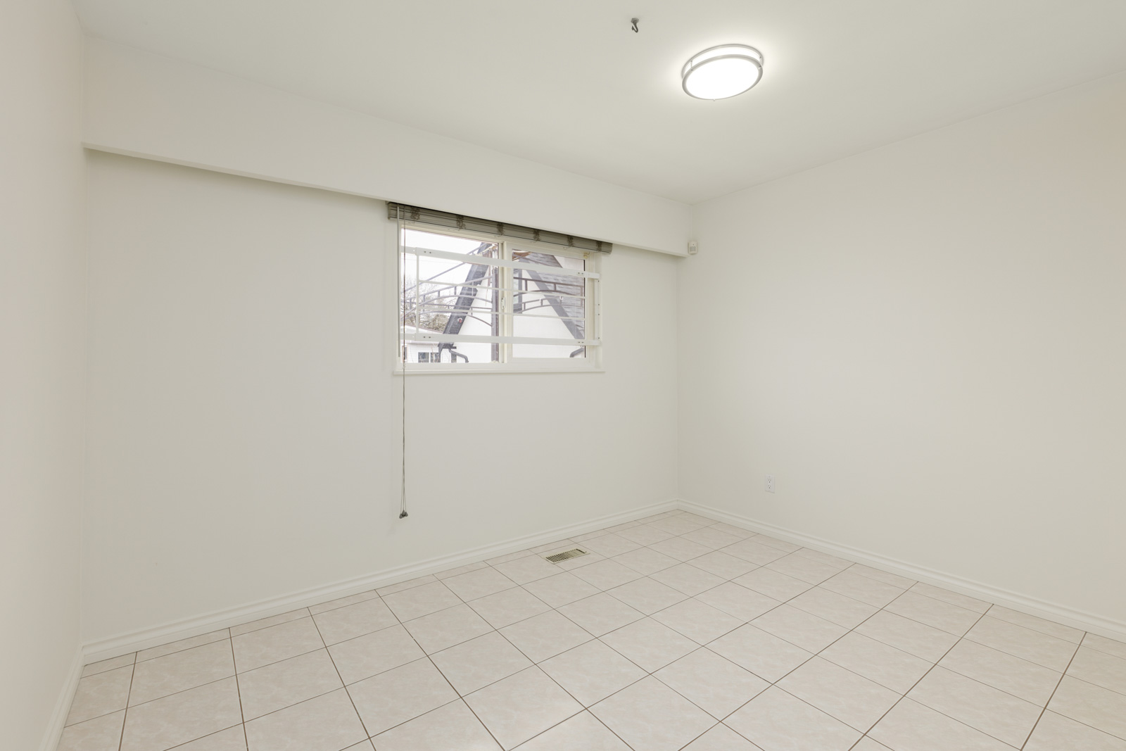 Empty room with white walls, tiled floor, a window with blinds partially open, and a ceiling light.