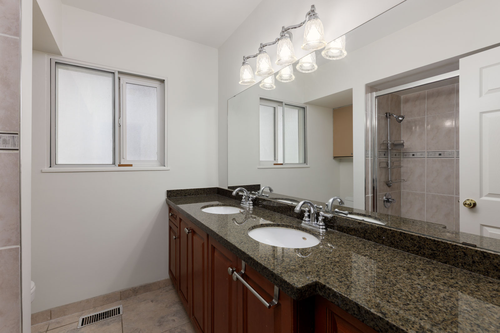 Bathroom with double sinks, granite countertop, wooden cabinets, large wall mirror, overhead lights, and a view of a walk-in shower and window.