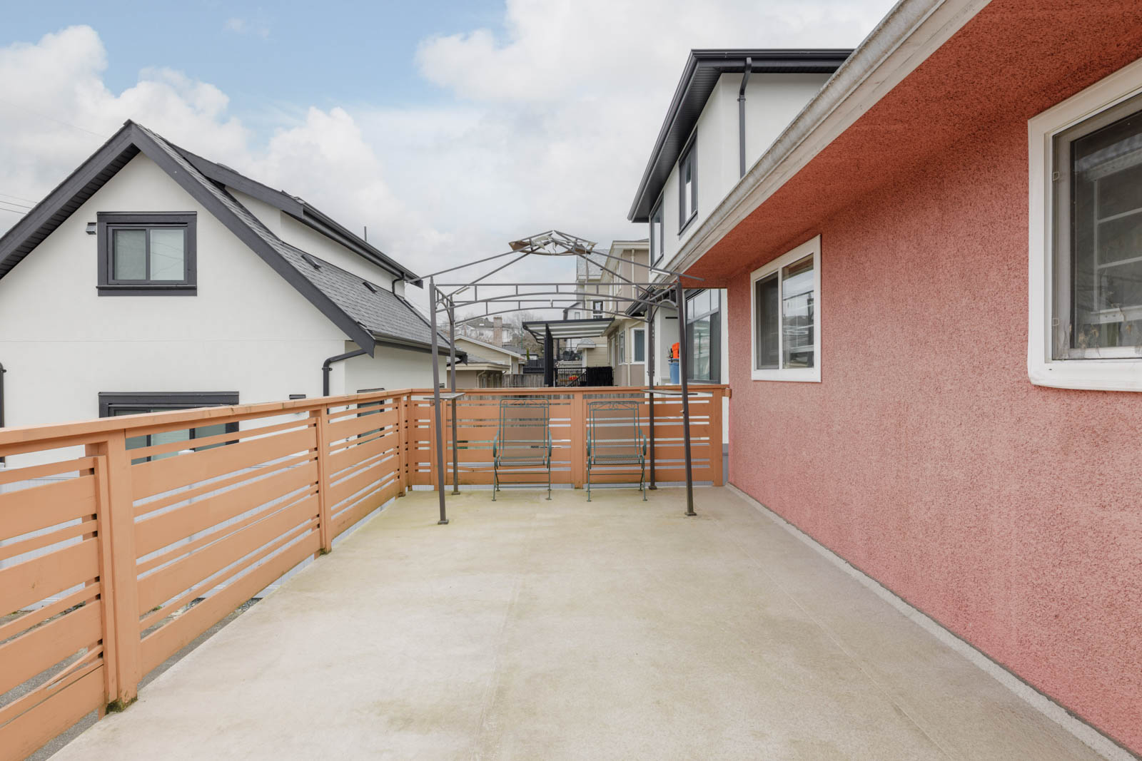 Spacious outdoor balcony with a peach-colored railing and wall, metal frame structure, and neighboring houses visible in the background under a partly cloudy sky.