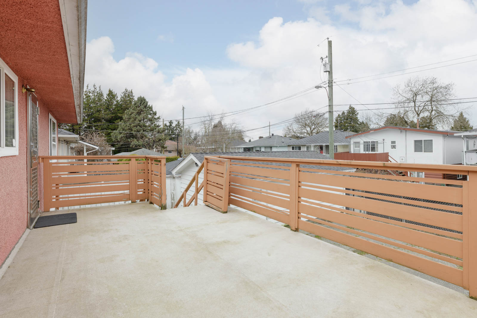 Spacious outdoor balcony with beige concrete floor, wooden railing, and view of neighboring houses and utility poles on a cloudy day.