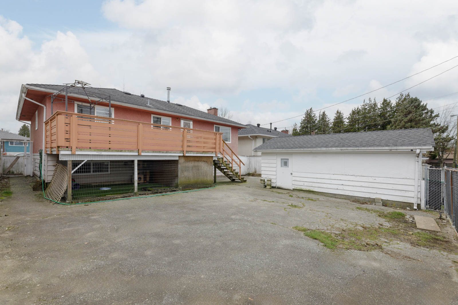 Back view of a house with a raised wooden deck, stairs leading to a paved backyard, and a detached white garage.