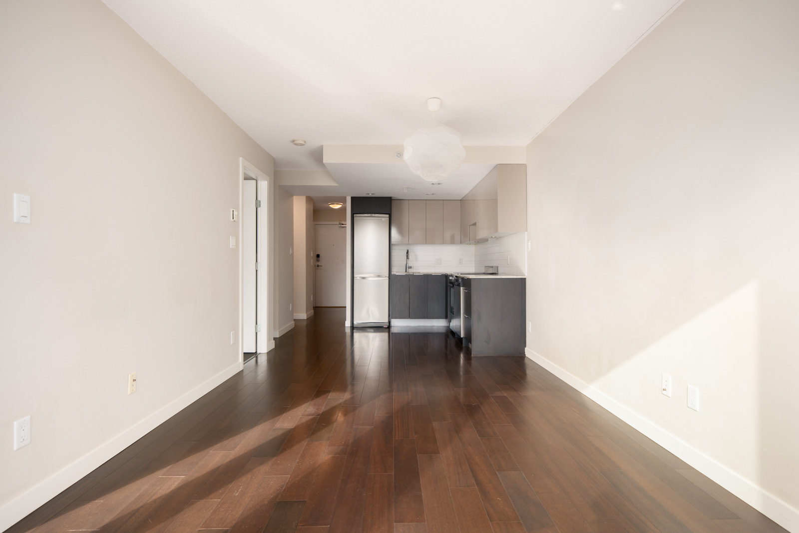 Unfurnished modern apartment with dark hardwood floors, light-colored walls, a small kitchen area, and stainless steel appliances. Natural light enters from the right.