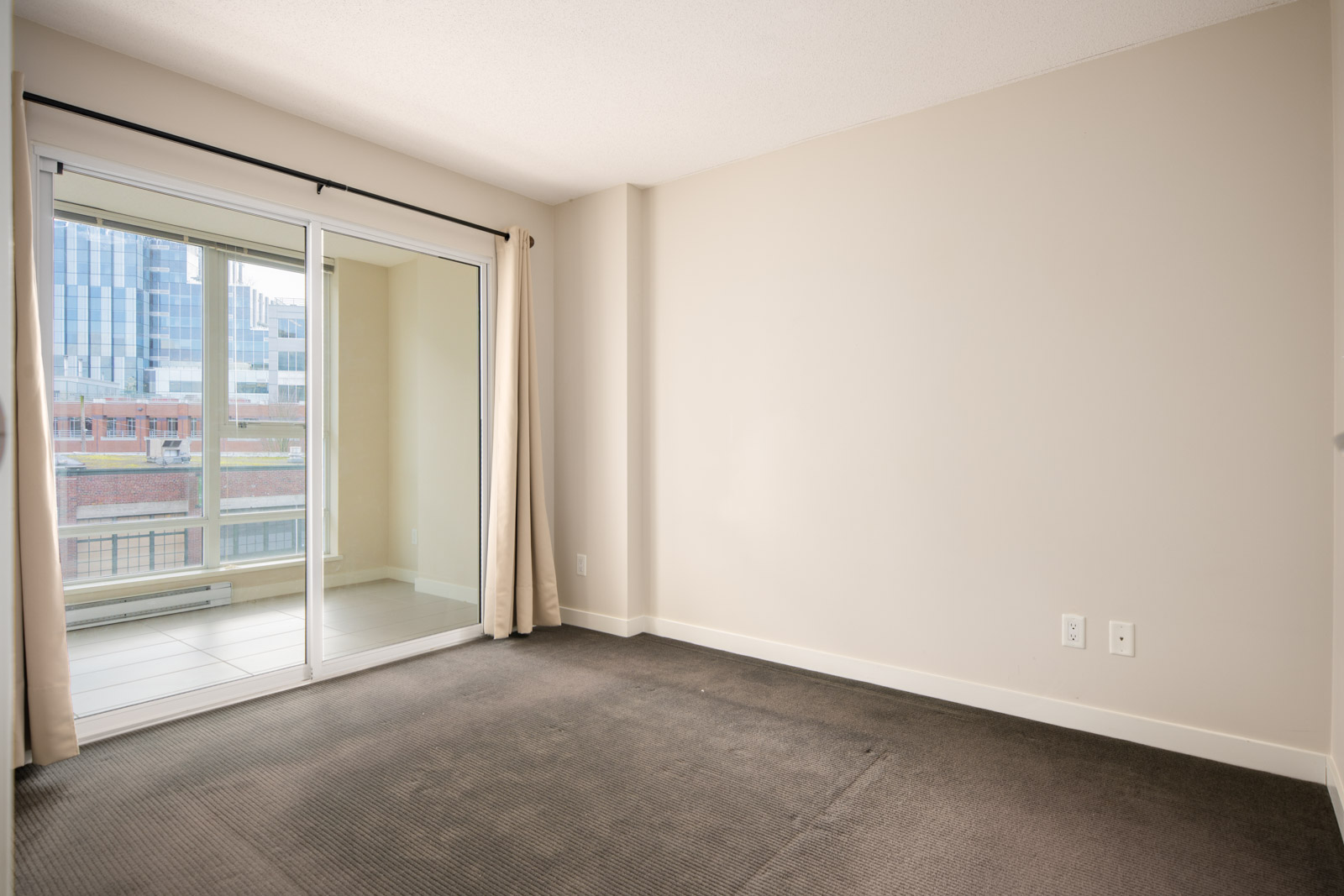 Unfurnished room with beige walls, dark carpet, and a large sliding glass door leading to an enclosed balcony with city buildings visible outside.