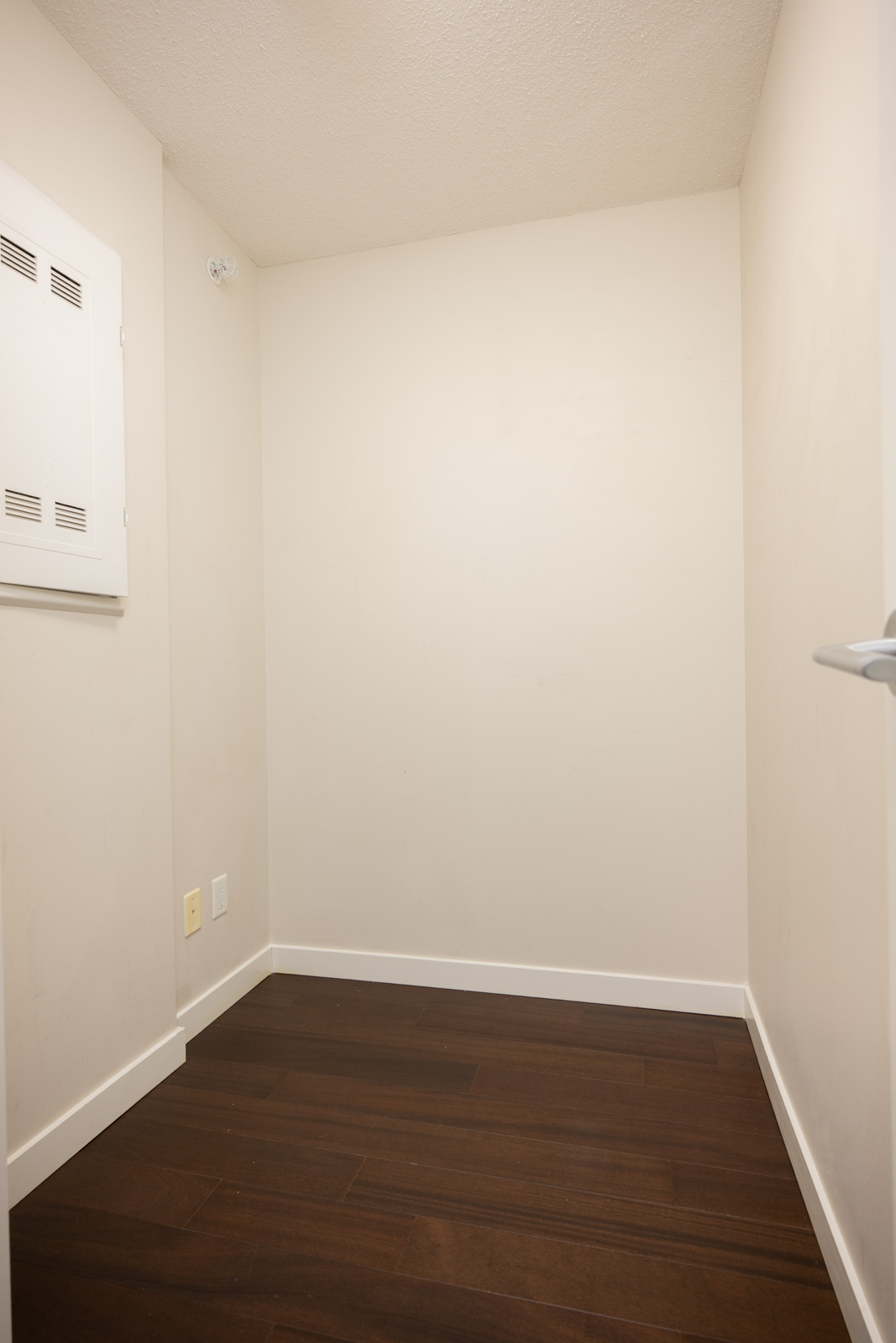 Small empty room with beige walls, dark wood floor, a white electrical panel, and a door partially visible on the right side.