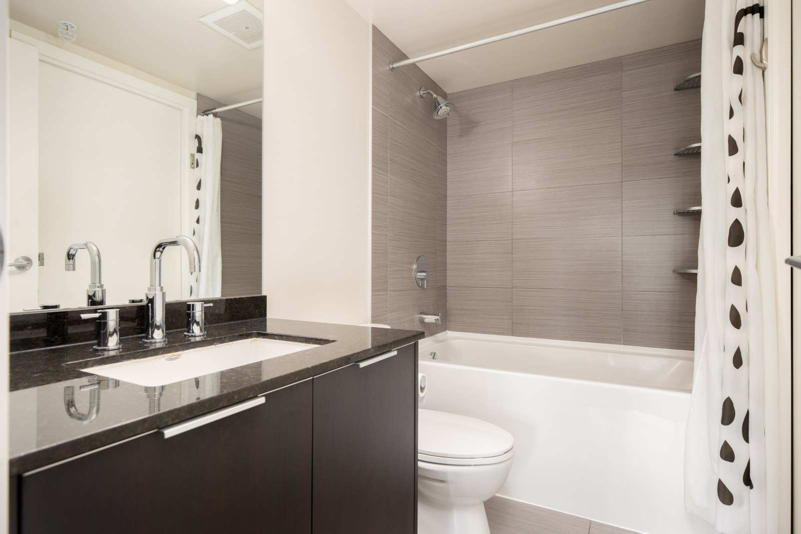 Modern bathroom with a dark wood vanity, white sink, chrome fixtures, a toilet, a bathtub with gray tile walls, and a white shower curtain with black dots.