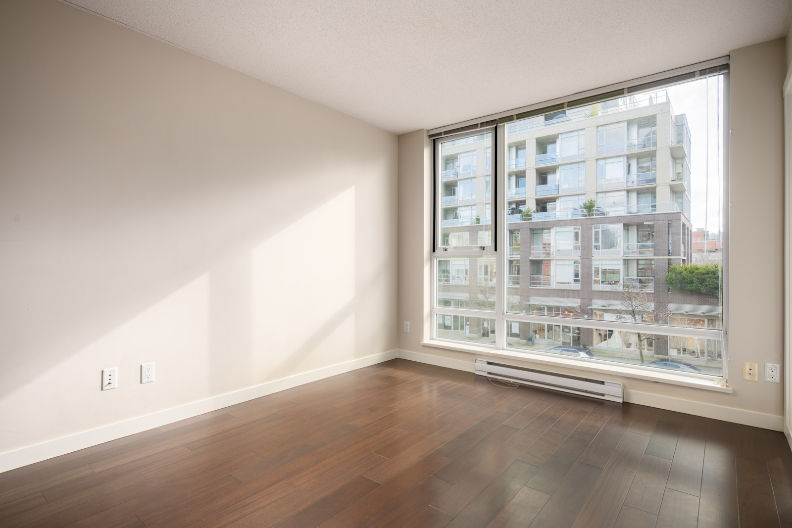 Empty room with light-colored walls, dark wood floor, large windows, and a view of a modern apartment building outside.