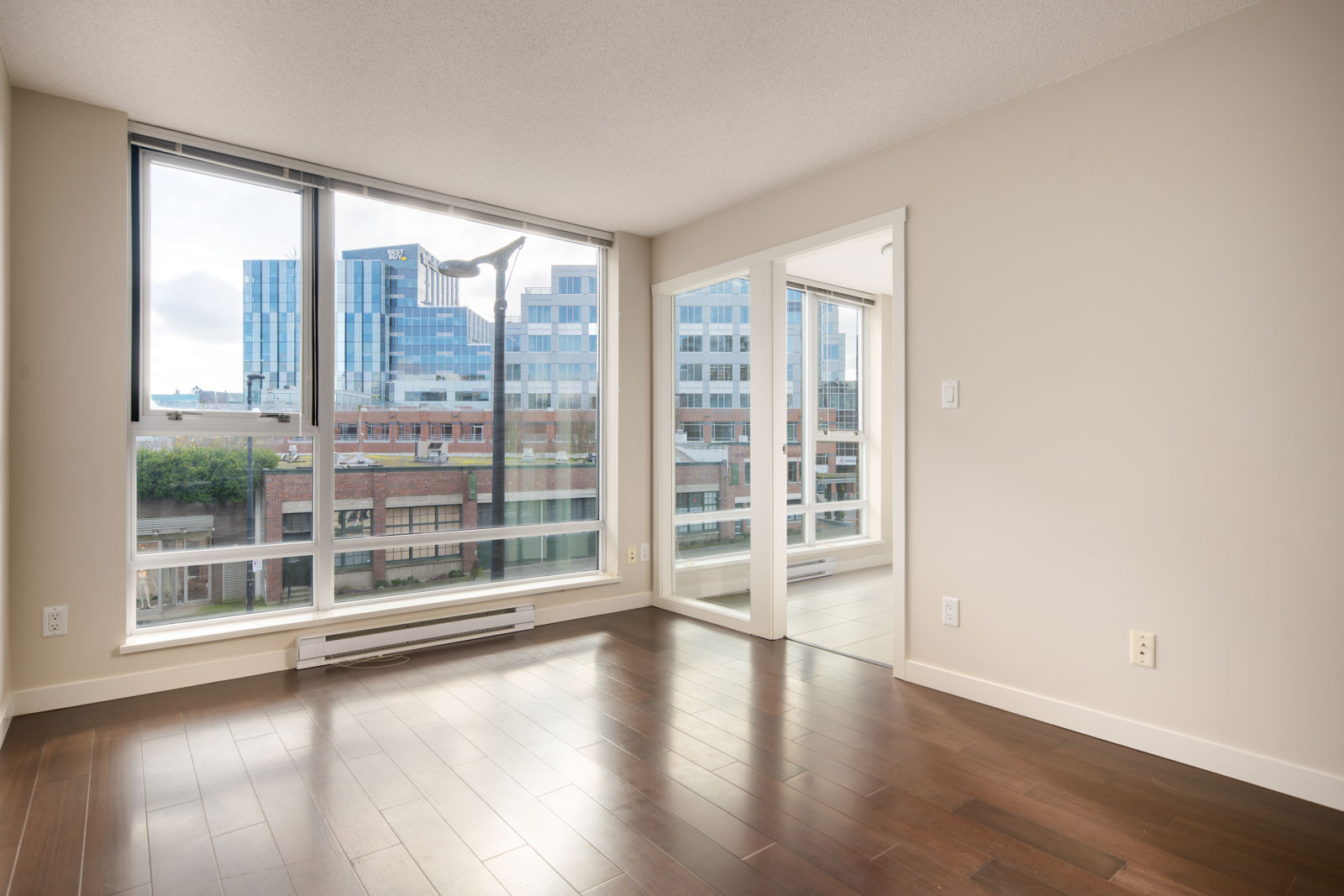 Unfurnished modern apartment room with large floor-to-ceiling windows, hardwood floors, and a view of city buildings outside.