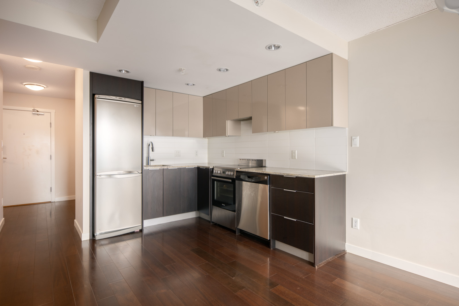 Modern kitchen with stainless steel appliances, dark wood flooring, and neutral cabinetry, adjacent to a light-colored wall and open entryway.
