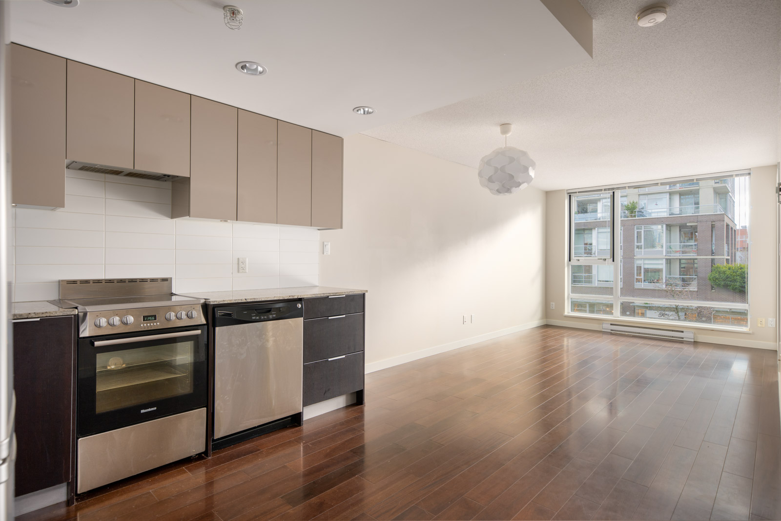 Unfurnished modern apartment with hardwood floors, an open kitchen featuring stainless steel appliances, and large windows overlooking nearby buildings.