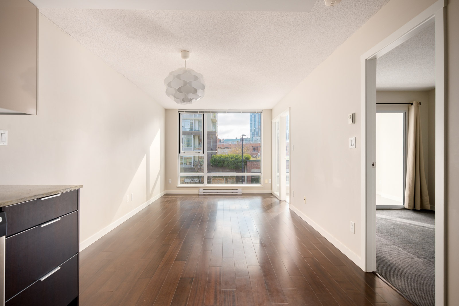 Bright, empty apartment room with dark hardwood floors, white walls, large window, modern light fixture, and open doorway leading to a carpeted room.