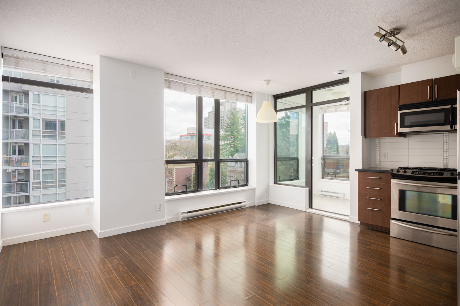 Bright, modern apartment with large windows, dark wood floors, white walls, and a kitchen area featuring dark cabinets, stainless steel appliances, and an over-the-range microwave.