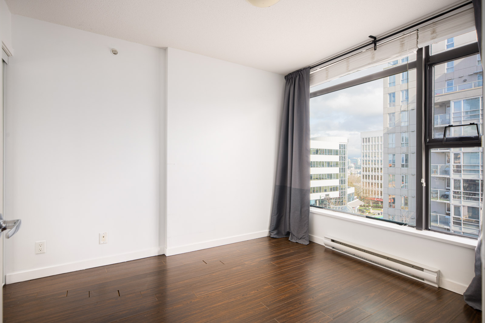 Empty room with white walls, dark wood floor, large window with gray curtains, and view of modern city buildings outside.