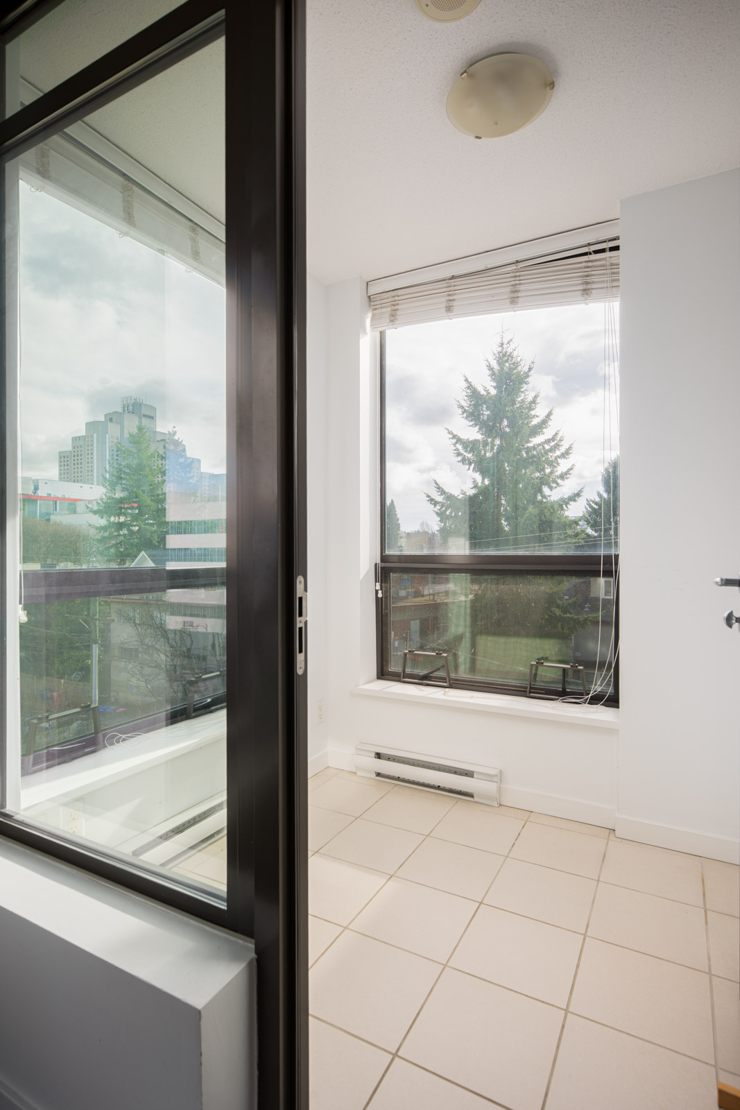 A small, tiled room with large windows and a sliding glass door opens to a balcony, offering views of trees and city buildings outside.