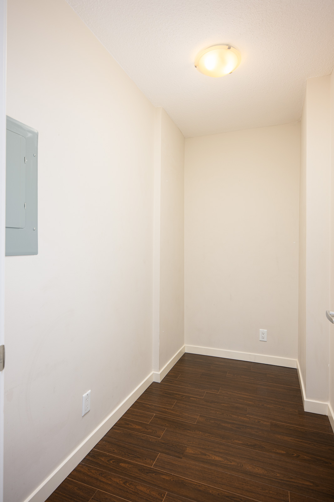 Small, empty room with beige walls, brown wood-look flooring, a ceiling light, electrical outlet, and an electrical panel on the left wall.