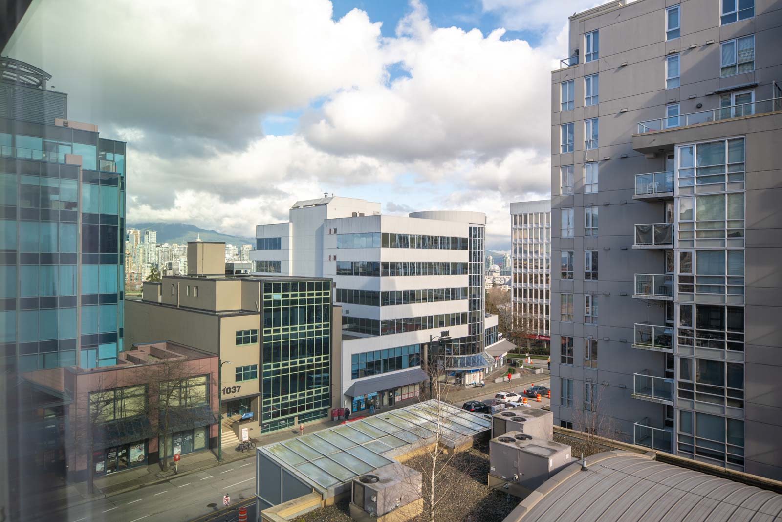 View of several modern office and apartment buildings in a city with a partly cloudy sky and a few pedestrians and vehicles on the street below.