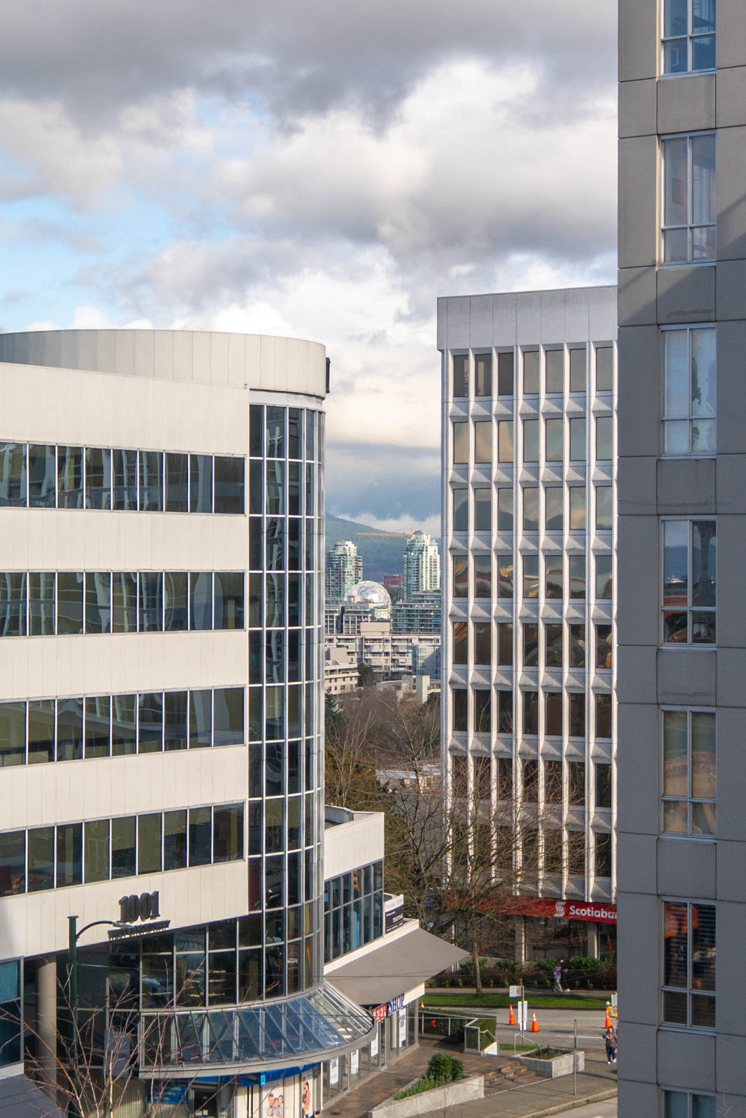 View of modern office buildings with glass windows in the foreground, cityscape and distant high-rises visible between them under a cloudy sky.