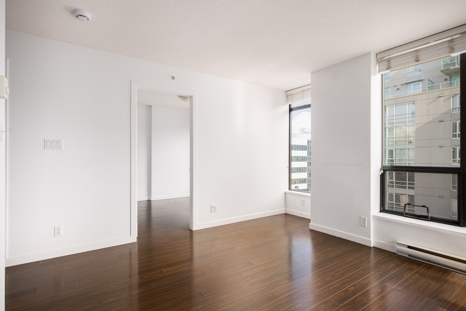 Empty, unfurnished apartment room with white walls, large windows, hardwood floors, and an open doorway leading to another room. Urban buildings are visible through the windows.