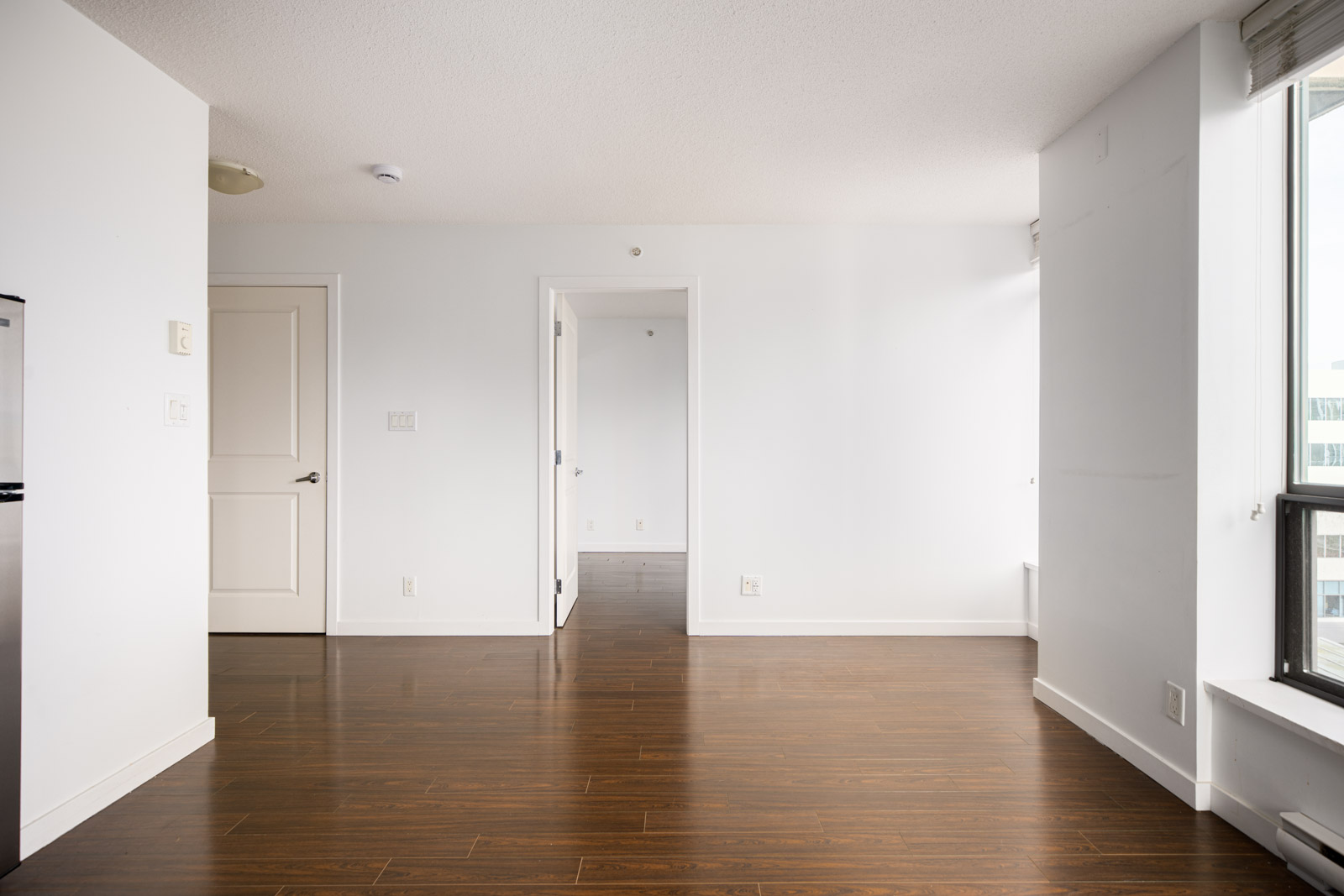 Empty apartment interior with white walls, dark wood floors, a window on the right, two doors, and visible natural light.