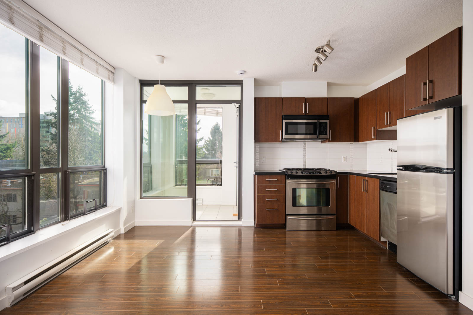Modern apartment kitchen with stainless steel appliances, brown cabinets, large windows, wood flooring, and an adjoining balcony.