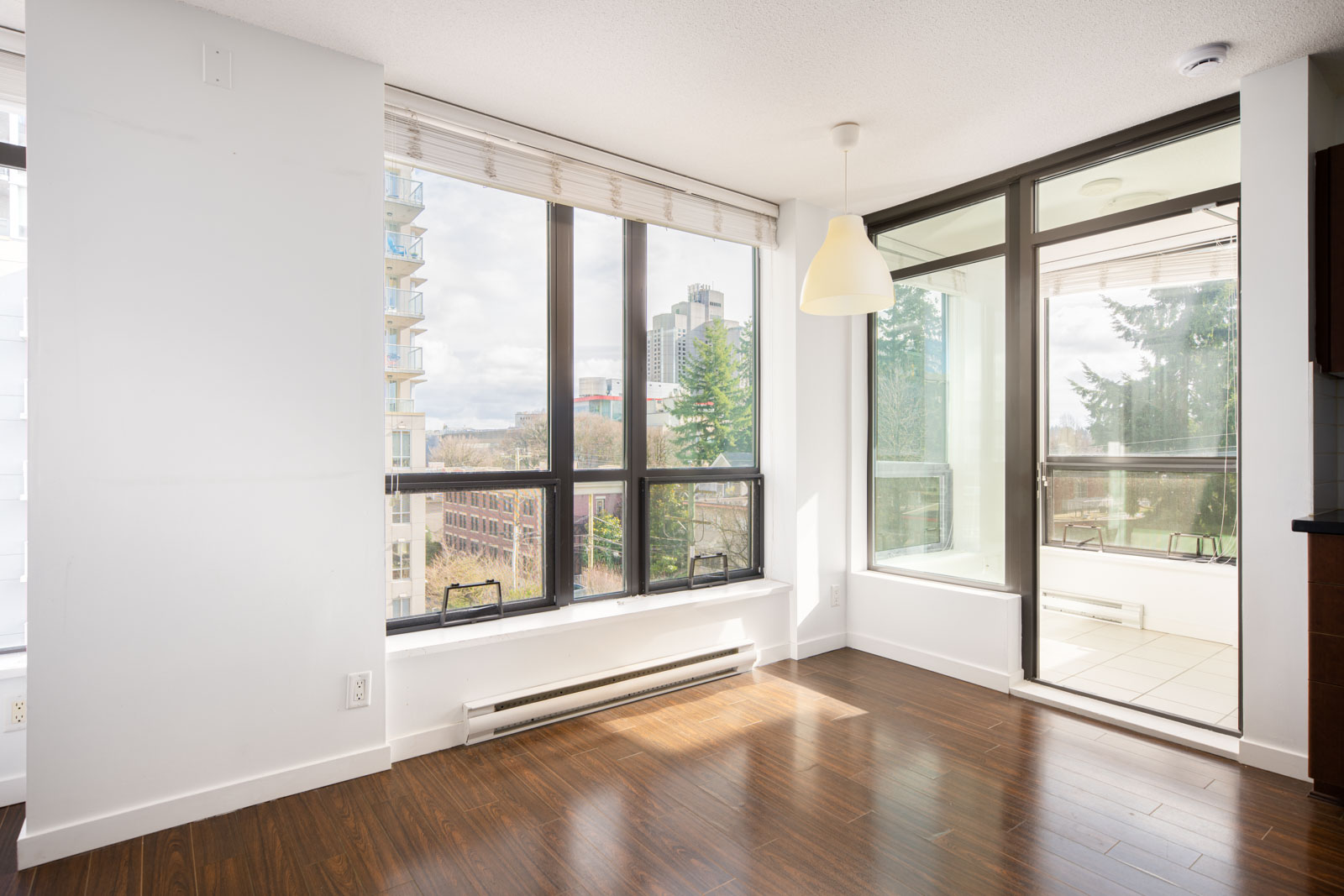 Bright, empty room with large windows, hardwood floors, a pendant light, and a glass door leading to a balcony with views of buildings and trees.