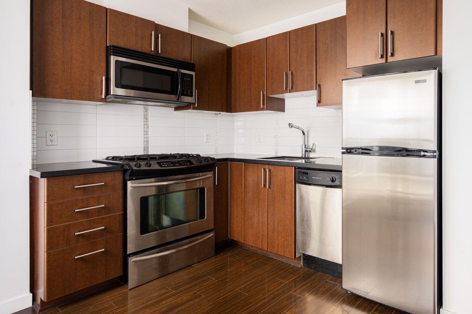 Modern kitchen with stainless steel appliances, brown wooden cabinets, black countertops, and white tile backsplash.